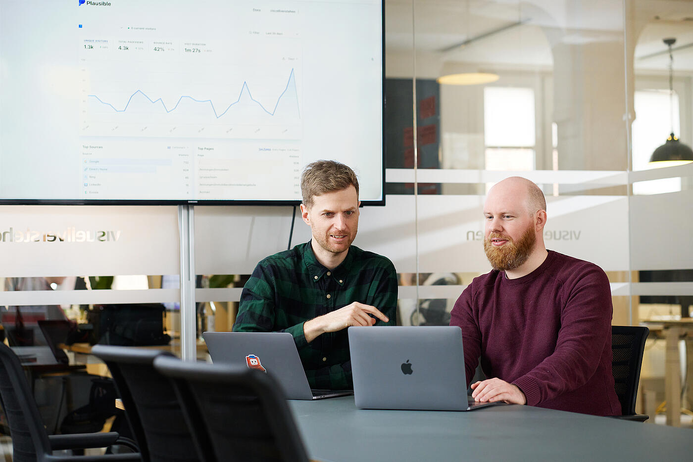 Zwei Mitarbeiter von visuellverstehen sitzen in einem Besprechungsraum vor zwei Laptops und sprechen über etwas.