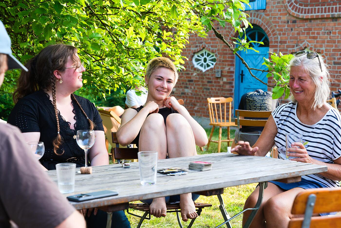 Mehrere Mitarbeiterinnen sitzen in idyllischer Kulisse an einem Tisch und lachen. Auf dem Tisch stehen Getränke und Spielkarten liegen bereit.