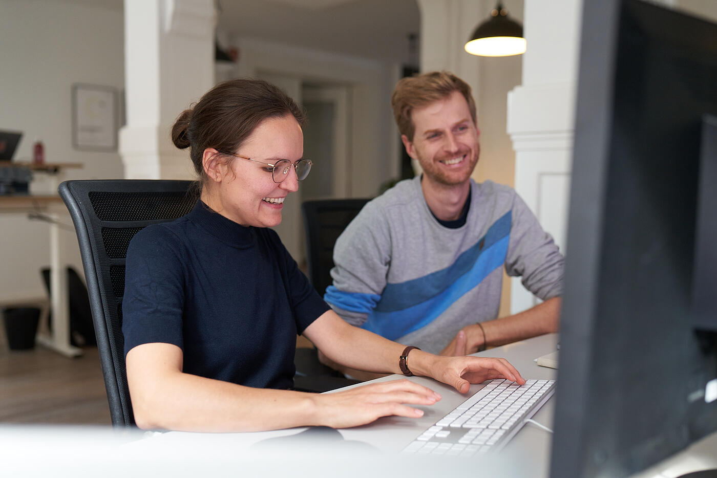 Zwei Mitarbeitende von visuellverstehen sitzen vorm Computer und lachen.