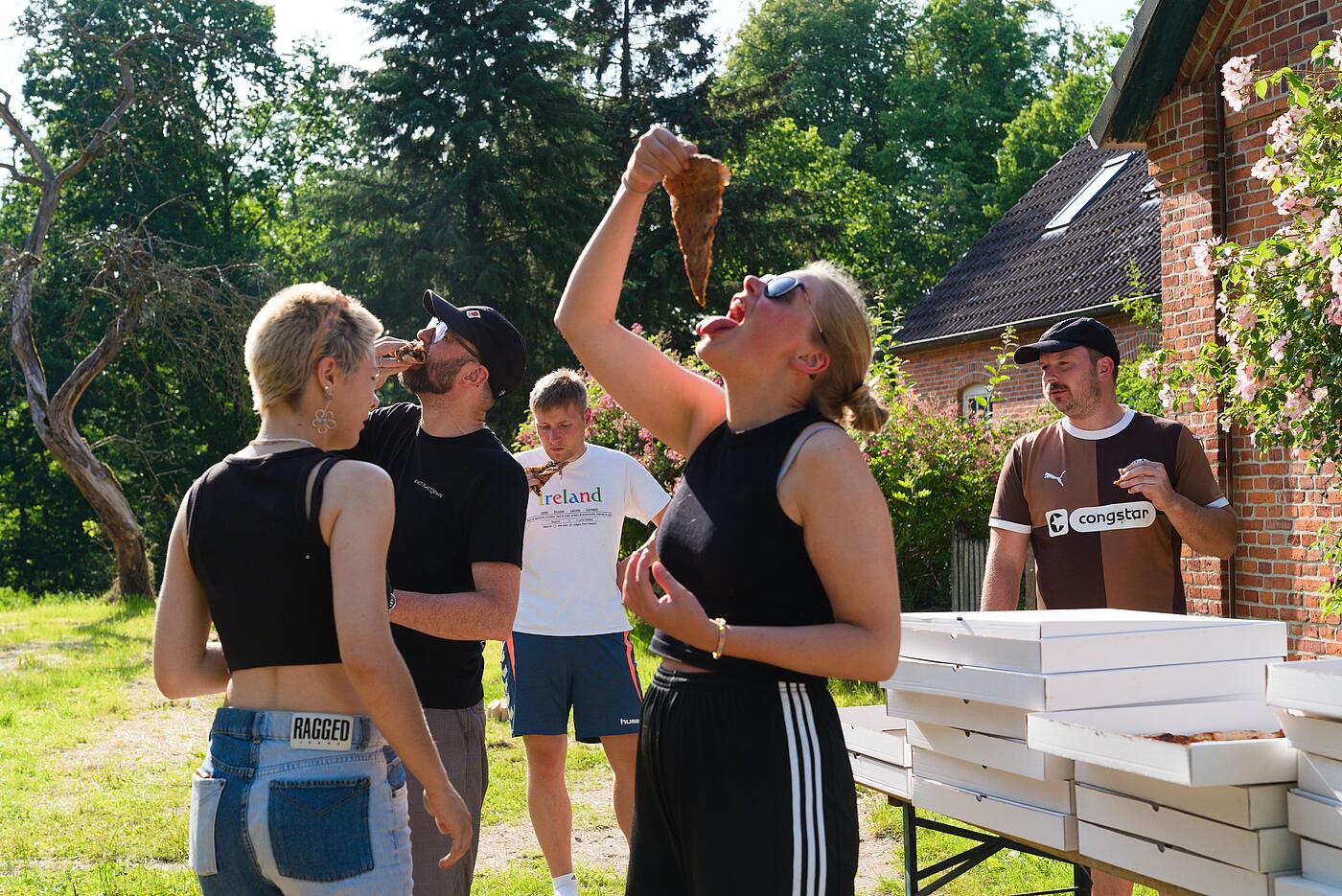 Teammitglieder stehen draußen in idyllischer Kulisse vor einem Tisch mit Pizzakartons. Im Vordergrund legt eine Mitarbeiterin den Kopf in den Nacken, um sich ein besonders großes Stück Pizza in den Mund fallen zu lassen.