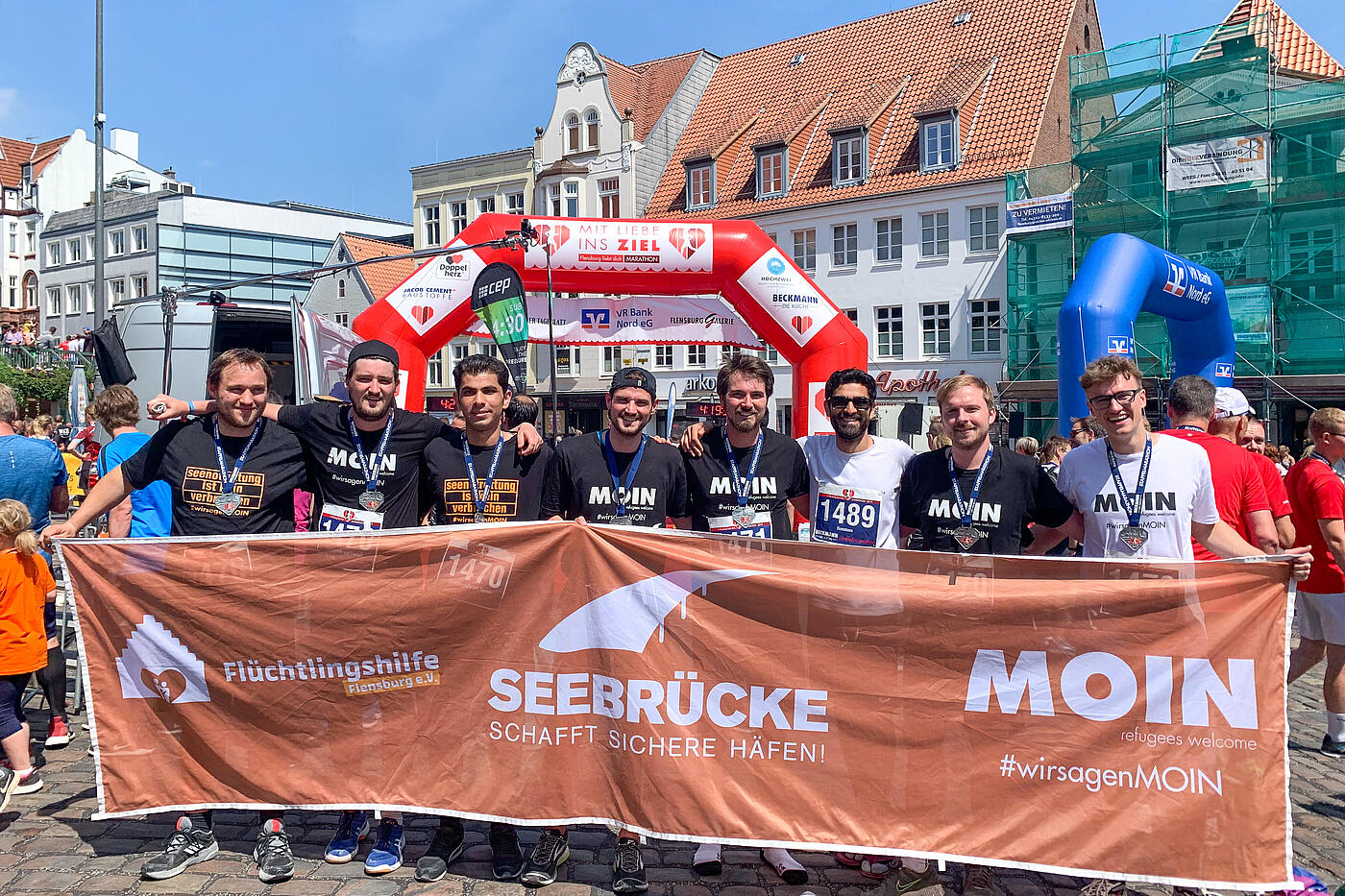 Eine Reihe von Männern verschiedener Nationen hält ein langes Banner mit der Aufschrift Seebrücke schafft sichere Häfen in die Höhe, im Hintergrund eine Hüpfburg.