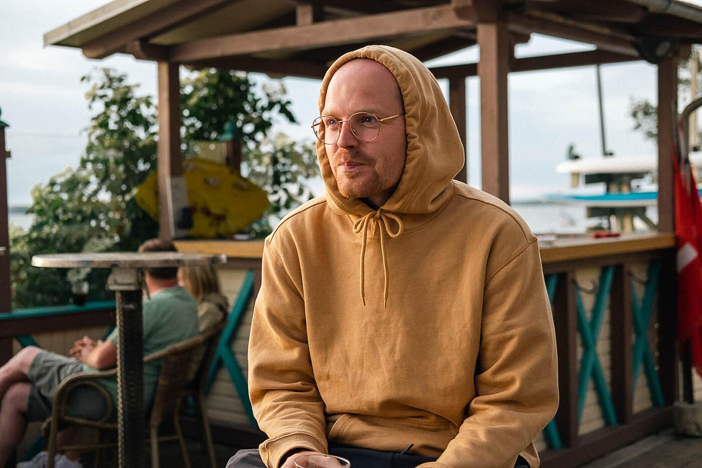 Ein Mitarbeiter von visuellverstehen sitzt im gelben Kapuzenpulli vor der Kamera, im Hintergrund eine Strandbar.