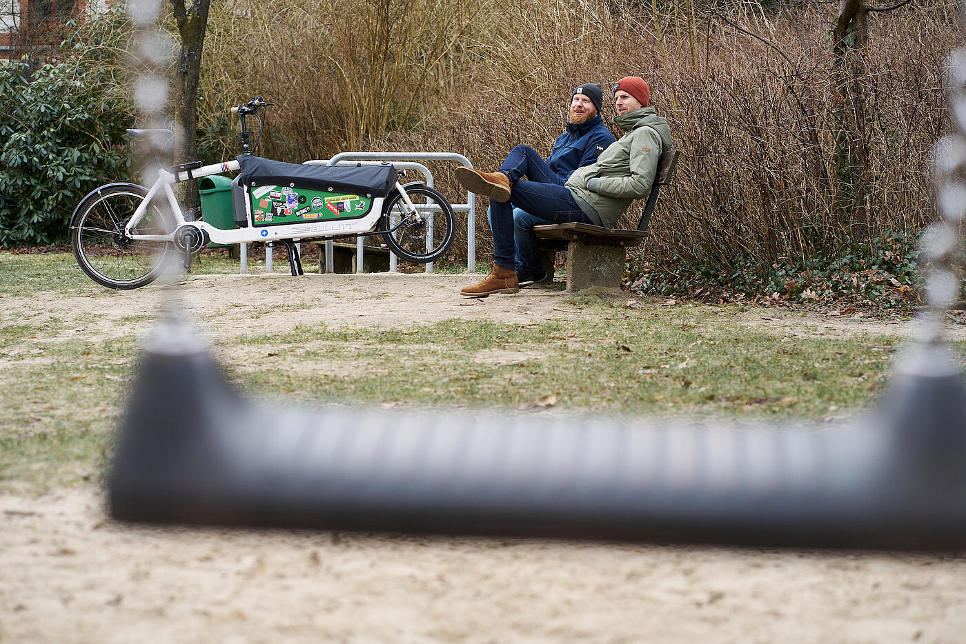 Zwei junge Väter aus dem Team von visuellverstehen sitzen warm eingepackt auf einer Bank am Rande eines Spielplatzes.