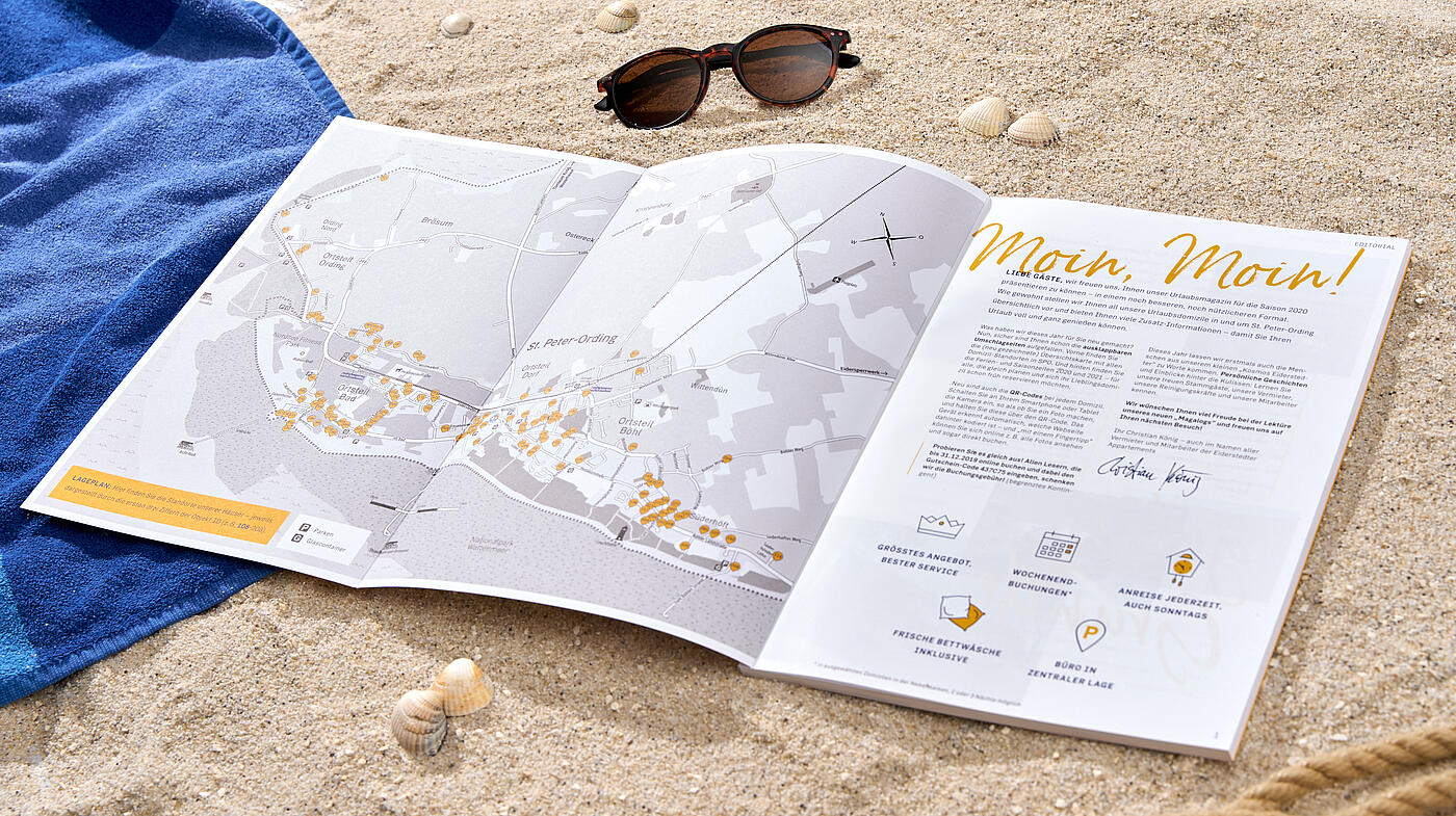 Eine aufgeschlagene Ausgabe des Eiderstedter-Magazins liegt am Strand zwischen einem Handtuch und einer Sonnenbrille.