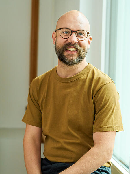 Jonas vor einem hellen Hintergrund. Er trägt dunklen Vollbart, Brille und ein hellbraunes T-Shirt. 