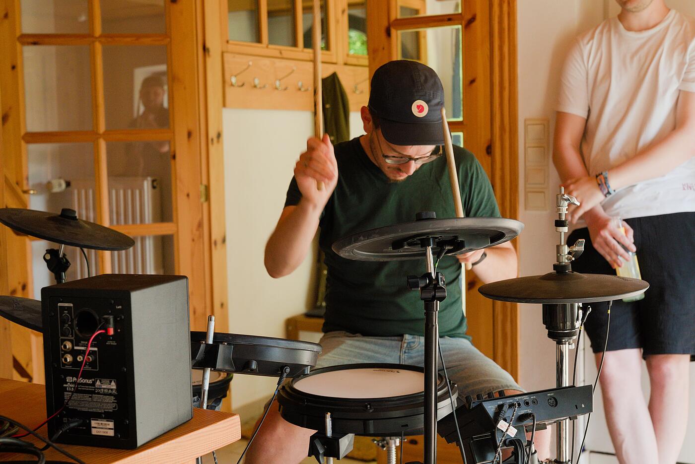 Ein Mitarbeiter spielt in einem warm erleuchteten Innenraum Schlagzeug an einem E-Drum-Set. Im Hintergrund lehnt ein weiterer Mitarbeiter an der Wand und hört zu.