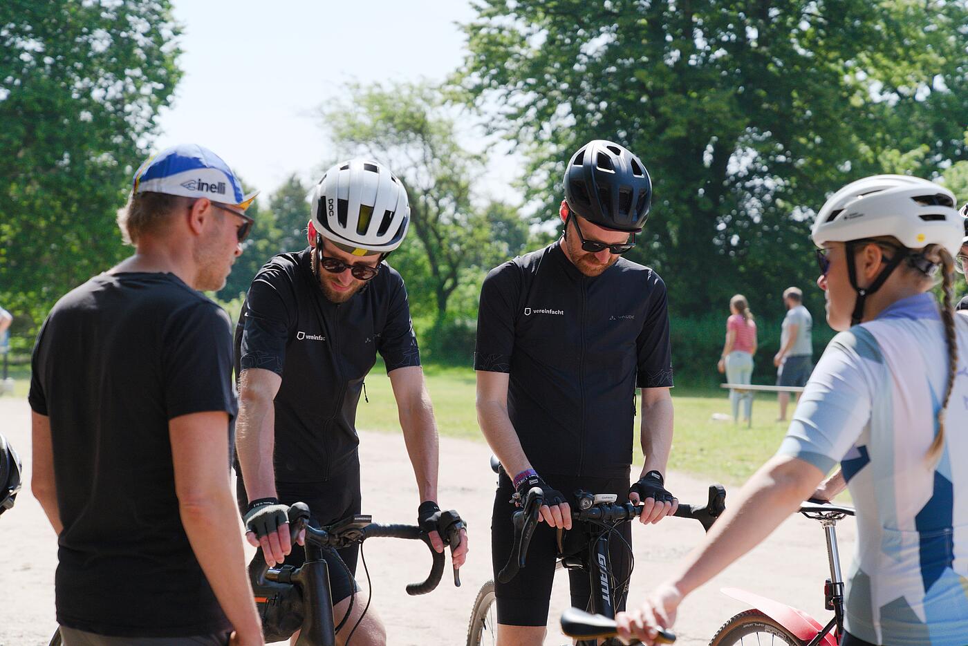 Mehrere Mitarbeitende in Rennsport-Kleidung und -Ausrüstung stehen mit ihren Fahrrädern auf einer Sandfläche, im Hintergrund Bäume und Büsche. Die Sonne scheint.