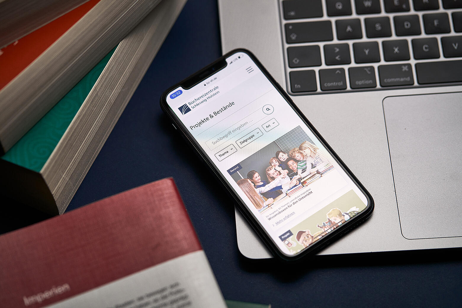 Ein Smartphone, das auf einem Macbook liegt, zeigt den neuen Internetauftritt der Büchereizentrale SH inmitten von Bücherstapeln auf einem Tisch.