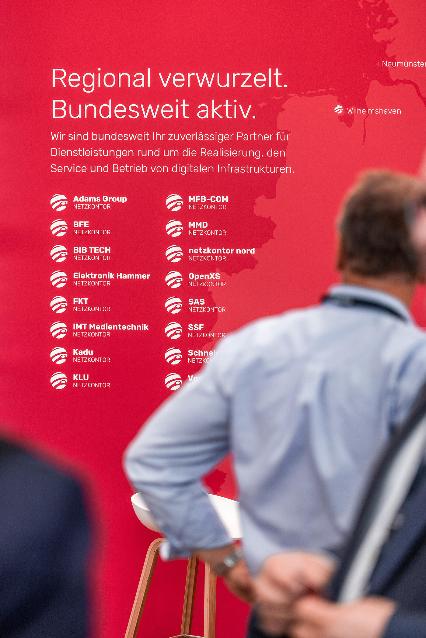 Ein rotes Plakat zählt in weißer Schrift auf einer Messe die verschiedenen Mitglieder des Netzkontors auf. Ein Mann steht davor und sieht sich das Plakat an.