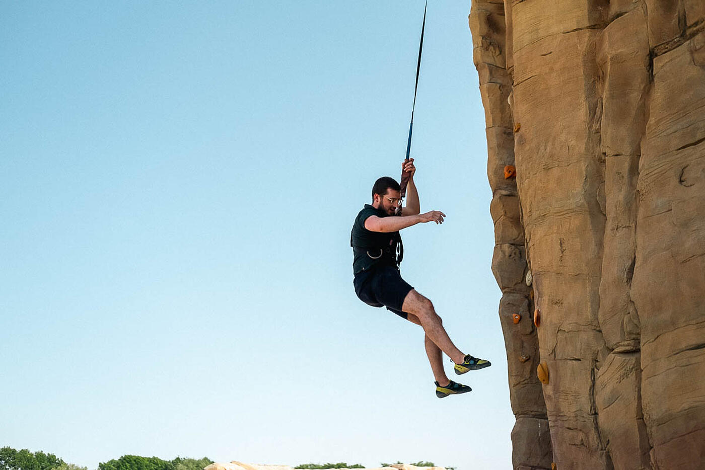 Ein Mitarbeiter von visuellverstehen hängt an einer Boulderwand vor blauem Himmel.