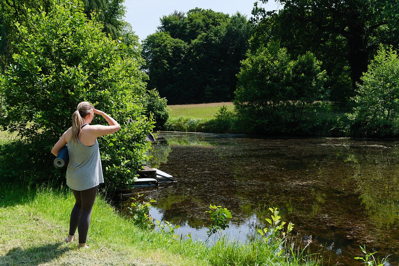 Eine Mitarbeiterin steht in Sportklamotten vor einem See im Grünen und schirmt die Augen gegen die Sonne ab. Sie hat eine Yogamatte unter dem Arm.