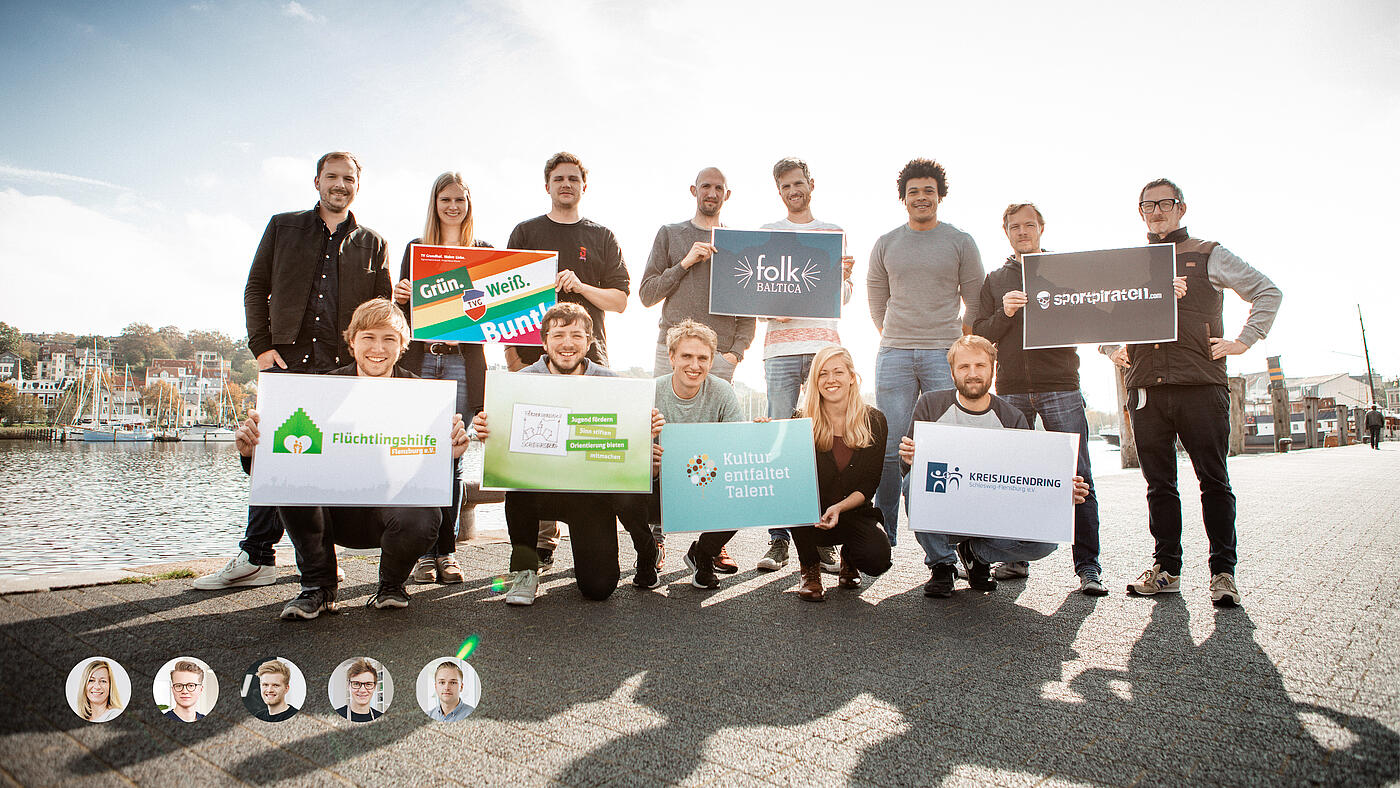 Das Team von visuellverstehen posiert am Flensburger Hafen mit den selbst kreierten Logos verschiedener Kund*innen.