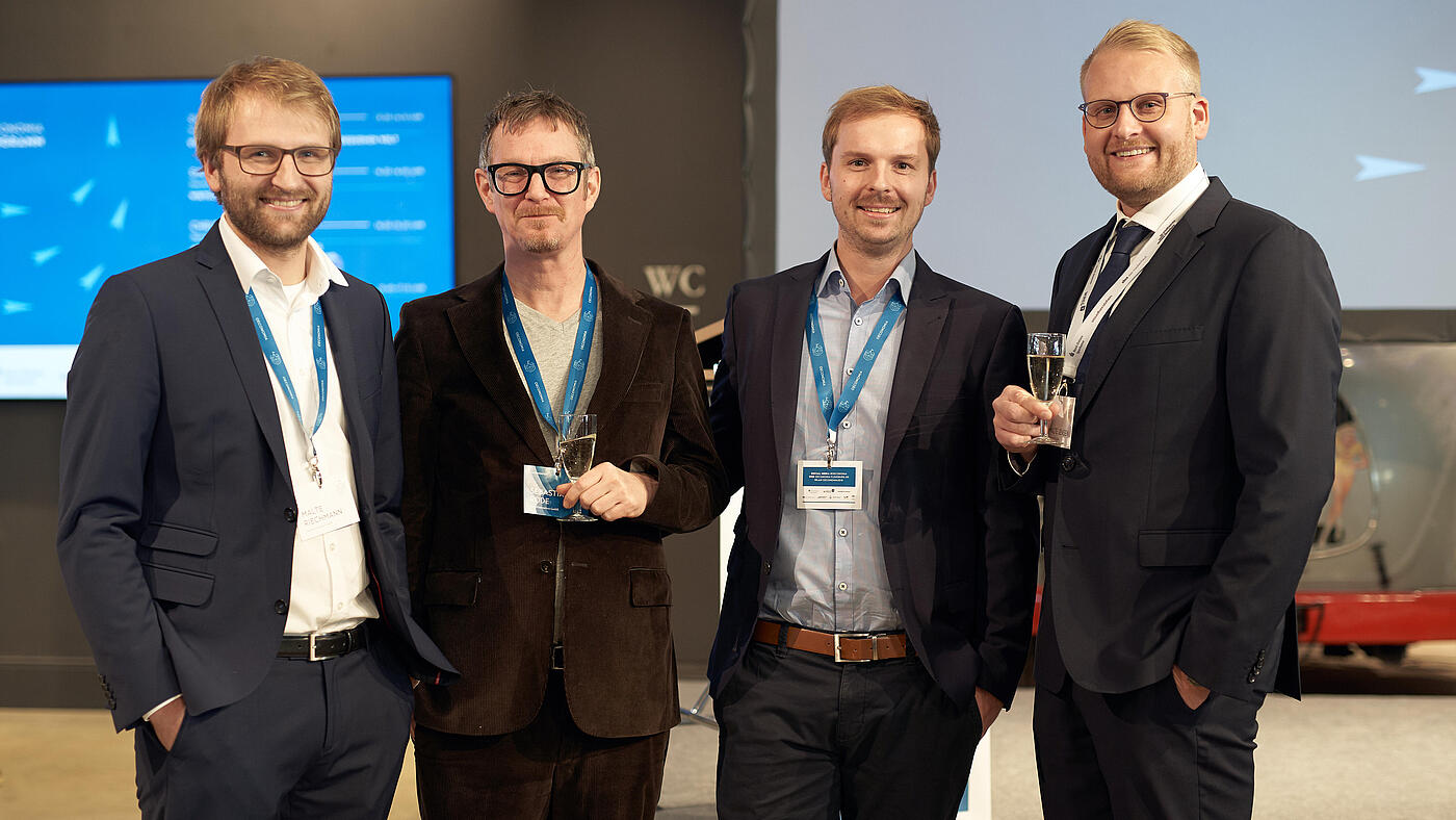 Die beiden Geschäftsführer und zwei Mitarbeiter von visuellverstehen posieren auf der Oeconomia 2018 mit Sektgläsern in der Hand für ein Foto.