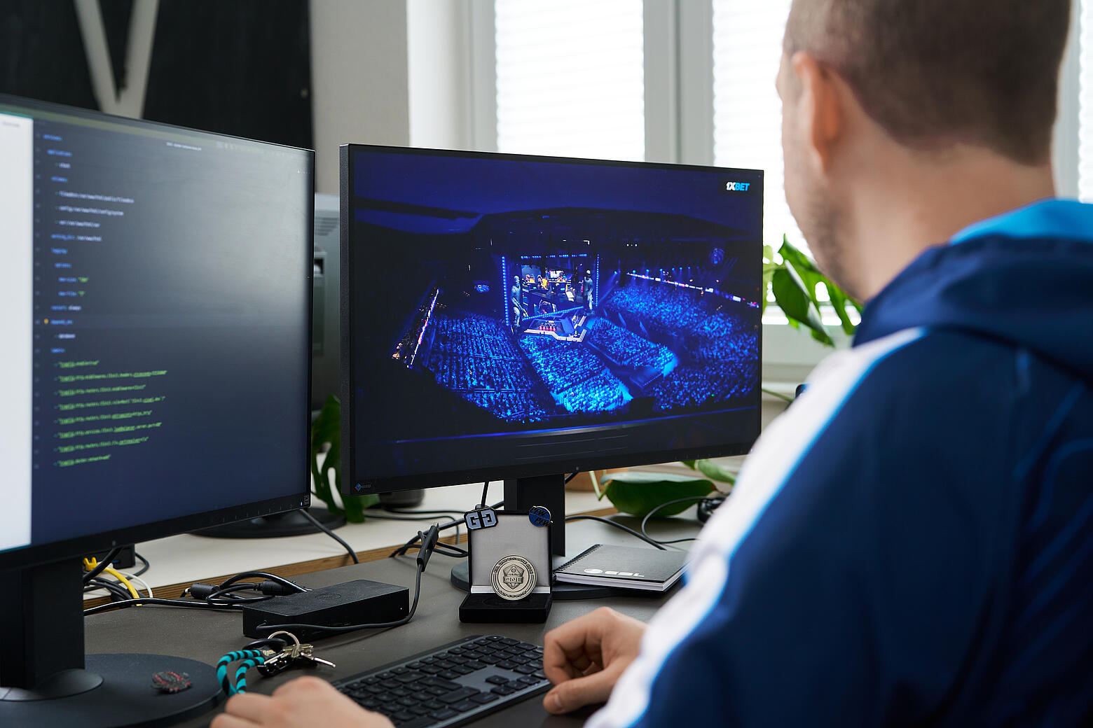 Blick über Philipps Schulter auf seine zwei Bildschirme. Während auf dem linken Monitor bunte Codezeilen zu sehen sind, wird auf dem rechten Monitor ein blau erleuchtetes E-Sport-Event in einem Stadium gestreamt.