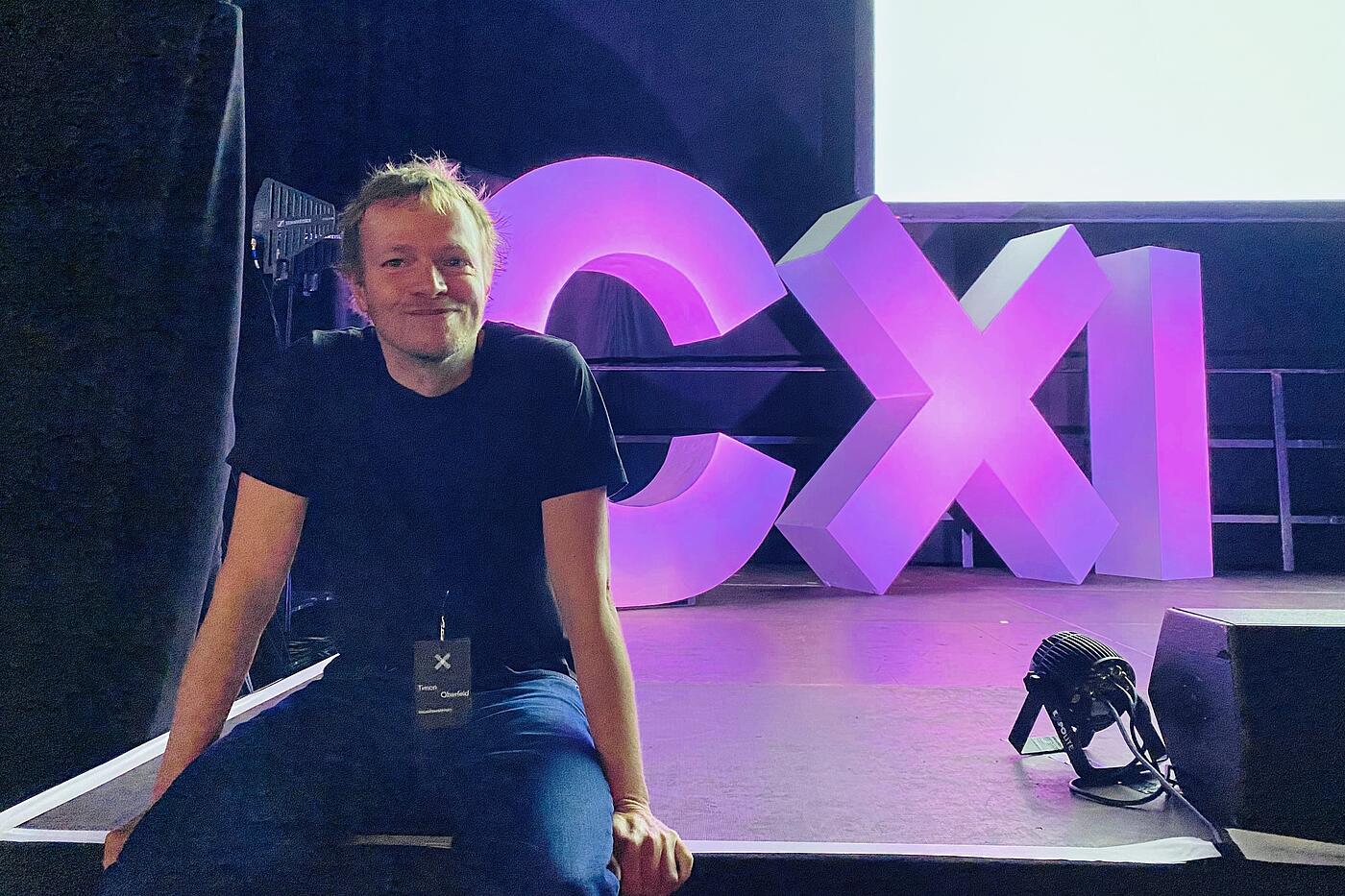 Ein Mitarbeiter von visuellverstehen sitzt auf einem Bühnenrand, im Hintergrund die großen CXI-Lettern.