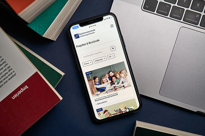 Ein Smartphone, das auf einem Macbook liegt, zeigt den neuen Internetauftritt der Büchereizentrale SH inmitten von Bücherstapeln auf einem Tisch.