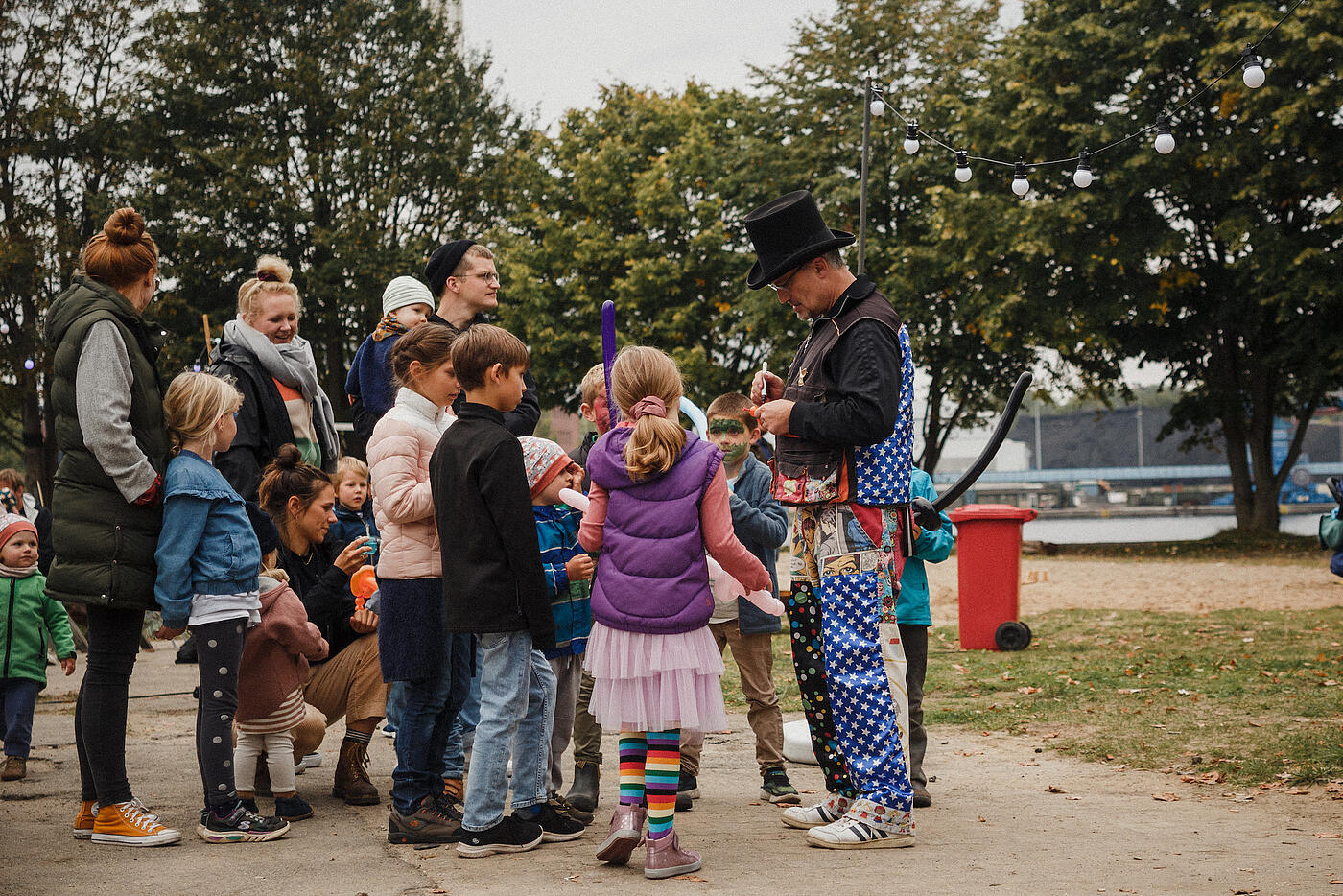 Ein von neugierigen Kindern und Eltern umringter Animateur mit Zylinder und bunter Kleidung knotet einen winzigen Luftballonhund.