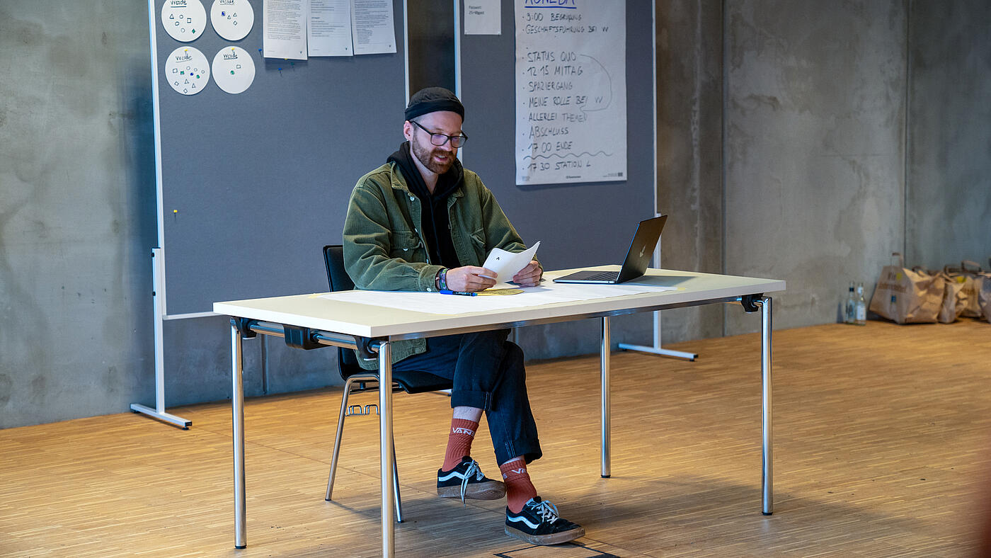 Ein Mitarbeiter von visuellverstehen sitzt ein einem Tisch vor mit Zetteln bespickten Pinnwänden und liest etwas vor. 