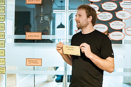 Ein Mitarbeiter von visuellverstehen hält einen Zettel mit der Aufschrift „Barrierefreie Websites“ in der Hand. Er steht vor einer Glaswand, auf der beschriftete Papiere befestigt sind.