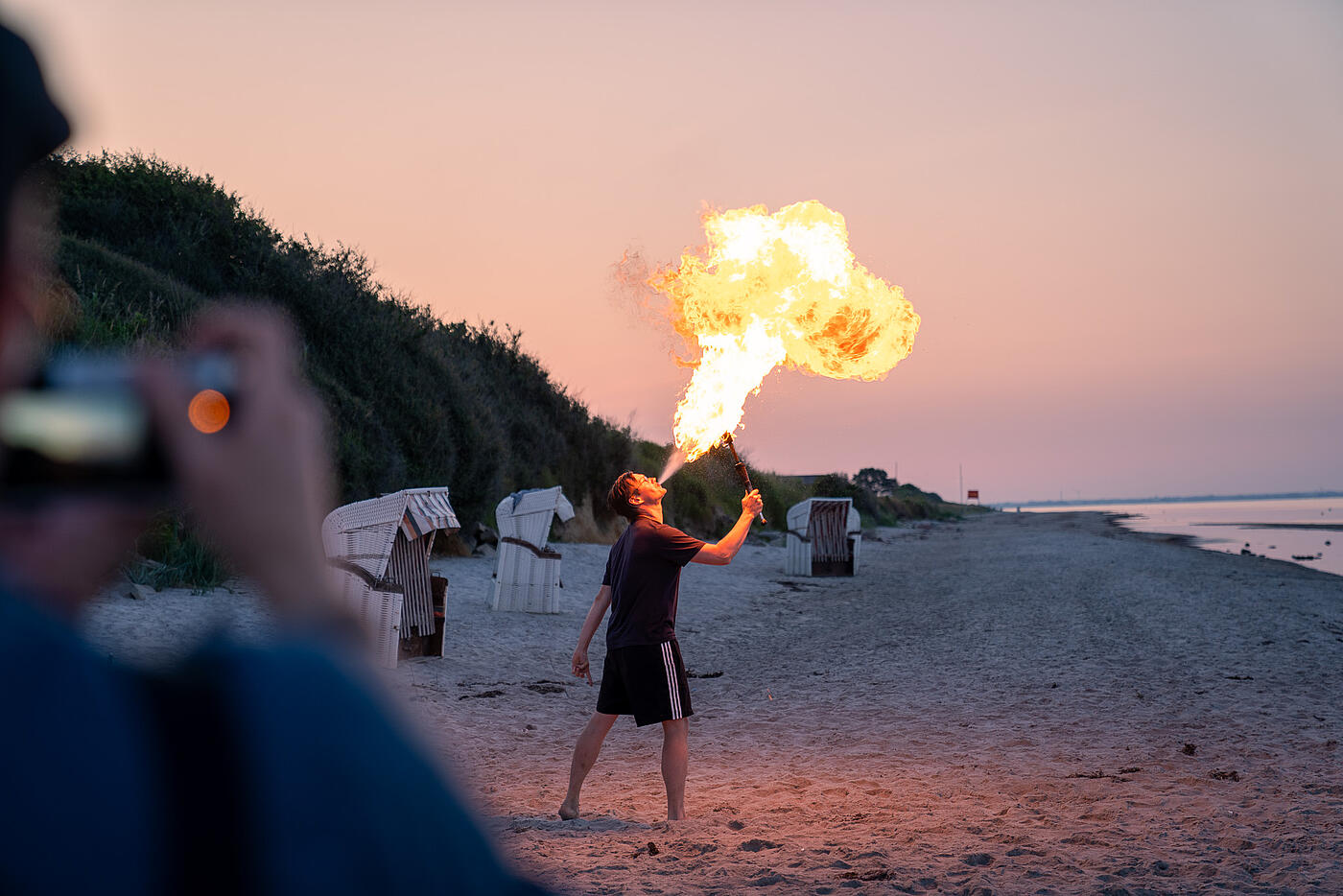 Ein einzelner Mitarbeiter von visuellverstehen übt sich im Feuerspucken, im Hintergrund der abendliche Strand.