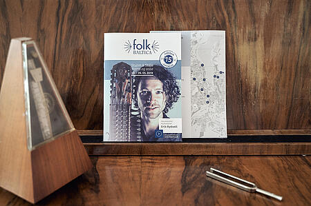 Ein Printprodukt für Folk Baltica steht auf einem Holztisch vor einem Holzhintergrund, im Vordergrund liegt eine metallene Stimmgabel.