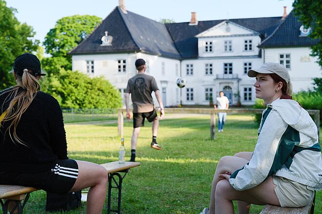 Vor einem malerischen historischen Gebäude spielen zwei Mitarbeiter Fußball, während zwei Mitarbeiterinnen im Vordergrund sitzen und sich umschauen.