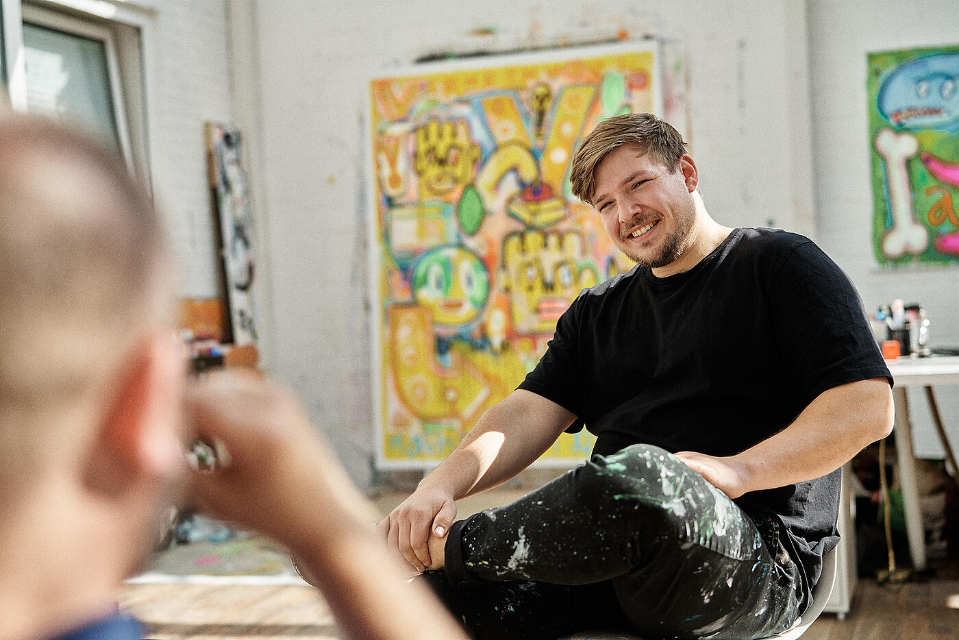 Ein junger Mann sitzt in einem Atelier vor dem Bild, das auch das Cover des Gemeinwohlmagazins zierte, und lächelt in die Kamera.