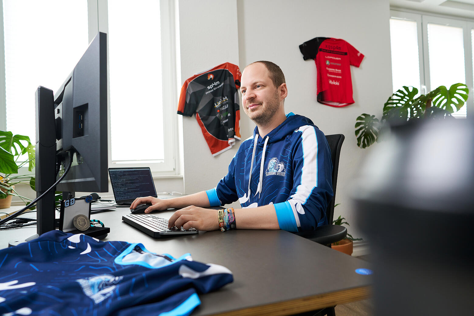 visuellverstehen-Mitarbeiter Philipp sitzt an seinem modernen, hellen Arbeitsplatz in der Agentur und lächelt. Im Hintergrund hängen zwei E-Sport-Trikots an der Wand, ein weiteres liegt neben Philipps Monitor auf dem Schreibtisch.