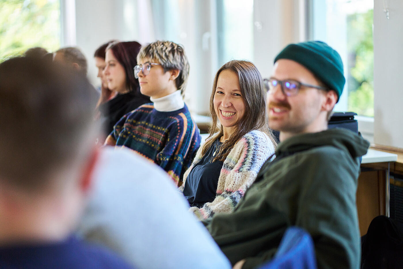 Menschen auf einer Stuhlreihe. Neben einem Mann mit Schnurrbart, Brille und Wollmütze sitzt eine Frau und lacht.