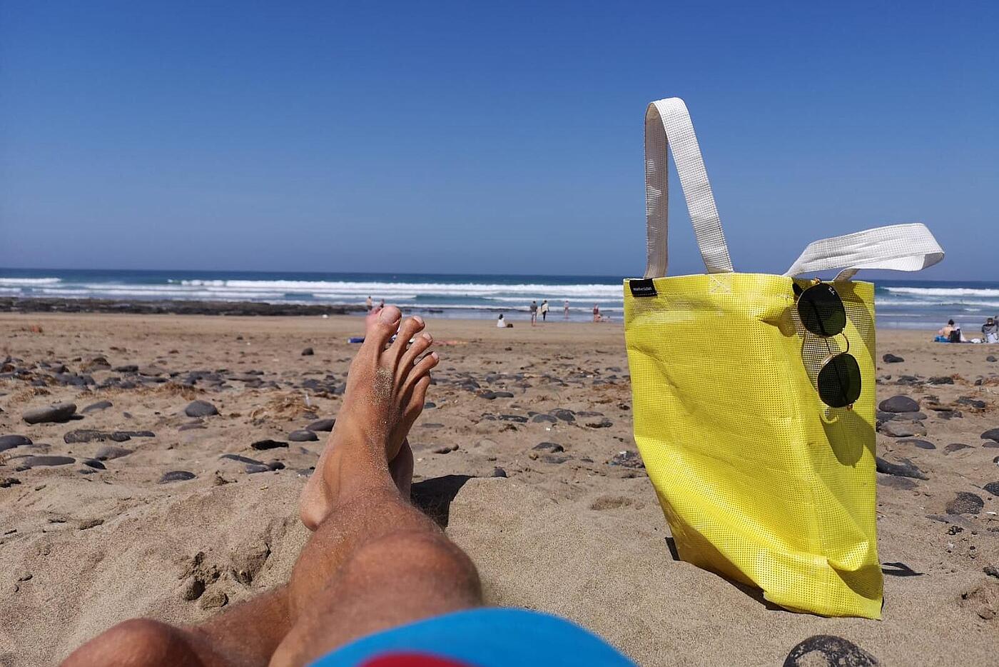 Ein Strand, im Vordergrund braungebrannte Beine und der gelbe Bannerbeutel von visuellverstehen.