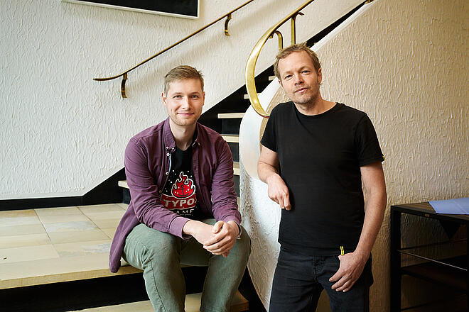 Rune und Timon an einem hellen Treppenaufgang. Rune sitzt auf der Treppe und trägt ein weinfarbenes, offenes Hemd und eine grüne Hose. Timon steht an das Geländer gelehnt. Er hat ein schwarzes T-Shirt und eine dunkelblaue Hose an.