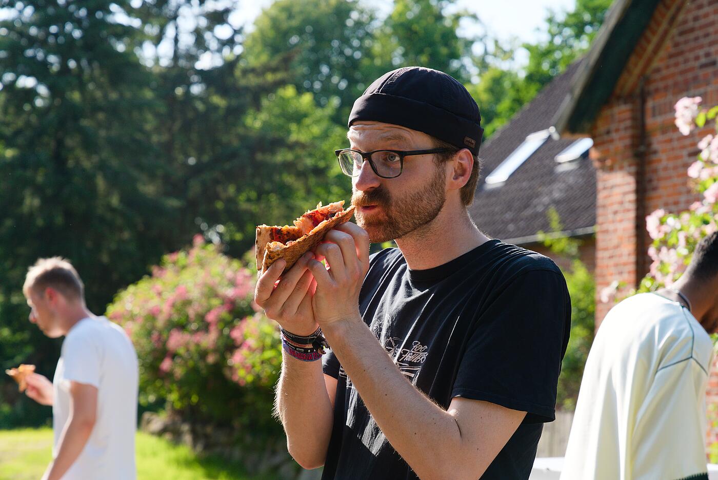 Ein Mitarbeiter hat ein Stück Pizza zusammengefaltet und beißt ab, im Hintergrund weitere Mitarbeitende und eine idyllische Kulisse mit viel Grün und einem historischen Gebäude.