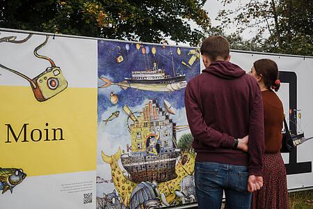 Auf dem Jubiläumsfest im Piratennest visualisiert eine lange Plakatwand die Historie des Unternehmens. Zwei Personen sehen sich diese aufmerksam an.
