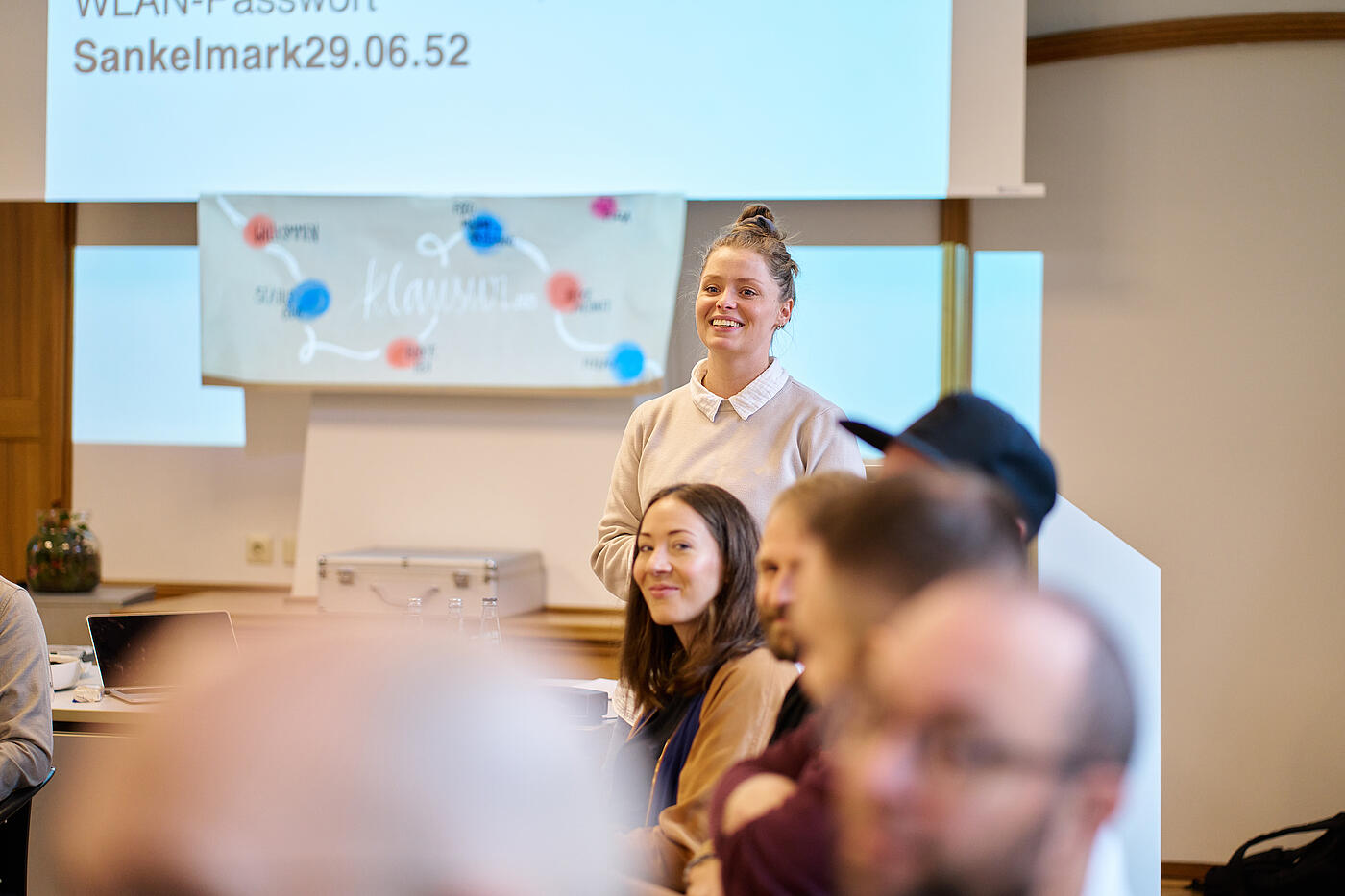 Lachende Frau mit hellen, hochgesteckten Haaren steht vor einer Gruppe Menschen.