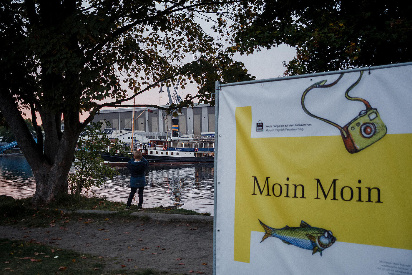 Während im Vordergrund ein Ausschnitt der Plakatwand zur Unternehmenshistorie zu sehen ist, steht im Hintergrund ein Junge am Wasser und sieht sich ein vorbeifahrendes Schiff an.