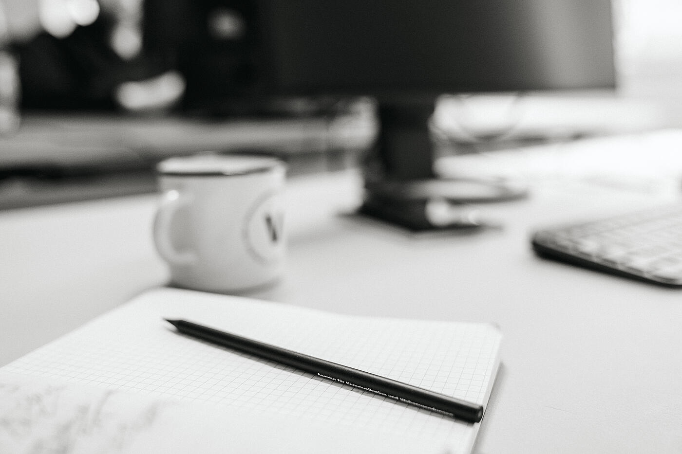 Ein Notizbuch mit einem Bleistift darauf, im Hintergrund ein visuellverstehen-Becher, in schwarzweiß.