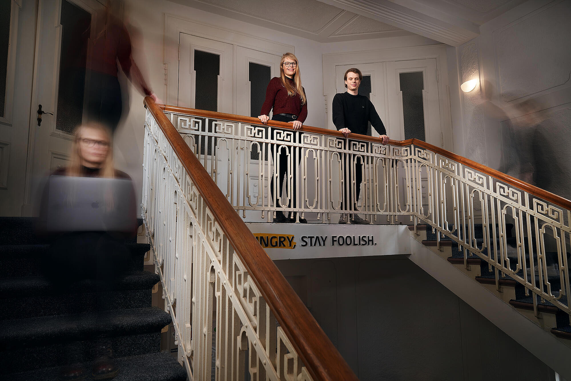 Aufnahme der beiden Werkstudierenden bei visuellverstehen, die im Treppenhaus selbstbewusst am Geländer lehnen, während im Zeitraffer aufgenommene Abbilder von ihnen die Treppen hoch und runter laufen.