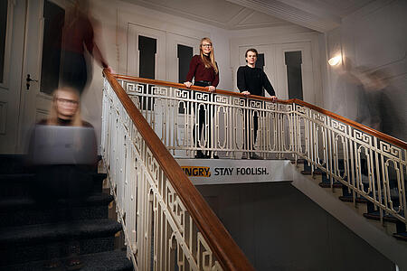 Aufnahme der beiden Werkstudierenden bei visuellverstehen, die im Treppenhaus selbstbewusst am Geländer lehnen, während im Zeitraffer aufgenommene Abbilder von ihnen die Treppen hoch und runter laufen.