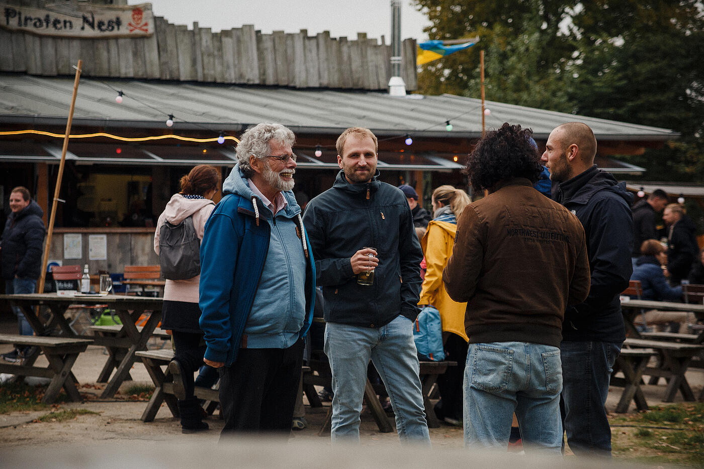 Mitarbeiter und Besucher auf dem Jubiläumsfest stehen mit Getränken in der Hand zusammen, im Hintergrund das Piratennest.