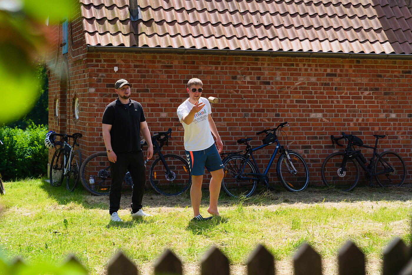 Rune steht auf einem Rasen und wirft einen Holzstab aus dem Wikingerschach Spiel. Er trägt Shorts, T-Shirt und Sonnenbrille. Neben ihm steht ein Mann, im Hintergrund lehnen Fahrräder an ein Gebäude aus rotem Backstein.