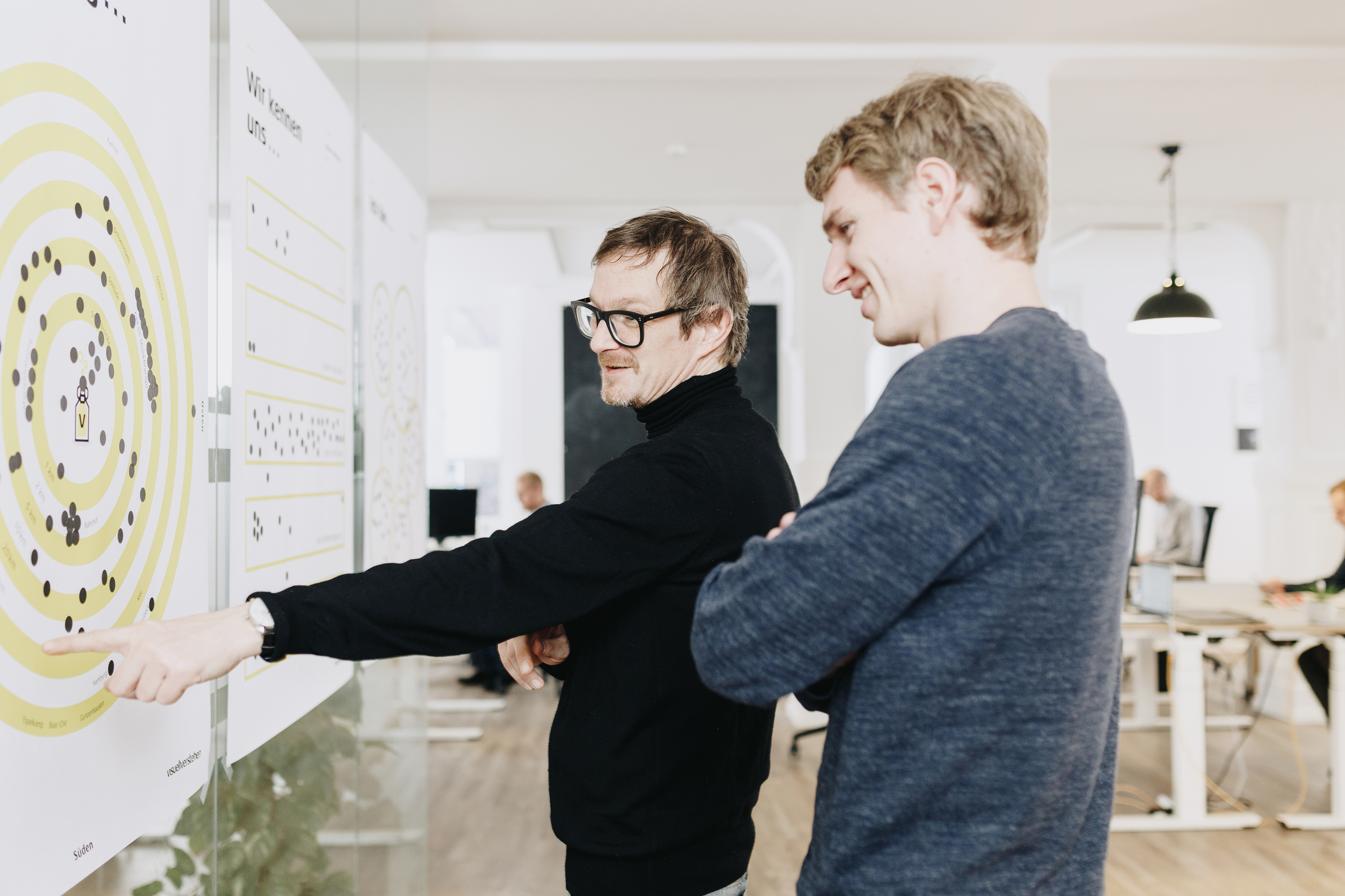 Zwei Mitarbeiter von visuellverstehen stehen vor einer Wand mit Haftnotizen, zeigen darauf und besprechen etwas.