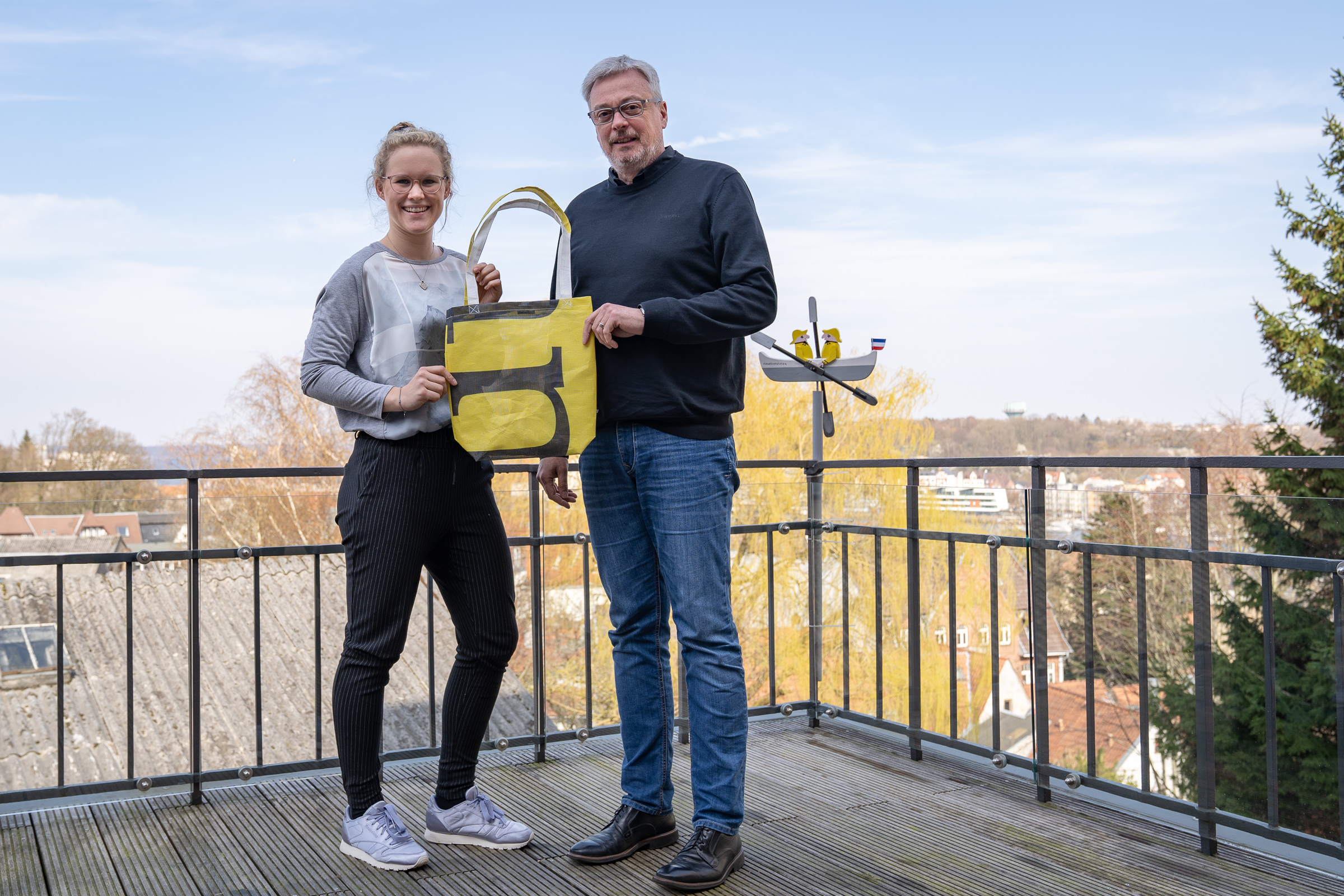 Eine Mitarbeiterin von visuellverstehen und der Vorstand der Mürwiker stehen auf der Dachterrasse und halten gemeinsam einen gelben Bannerbeutel von visuellverstehen hoch.