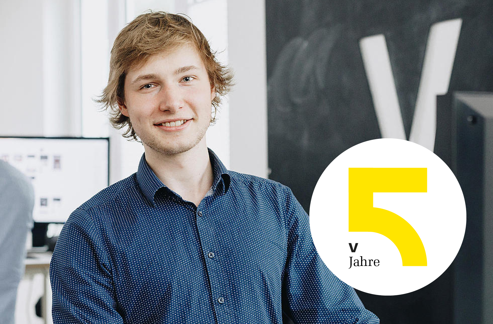 Porträtaufnahme eines Mitarbeiters bei visuellverstehen an seinem Arbeitsplatz, davor ein Badge mit einer gelben 5 darauf.