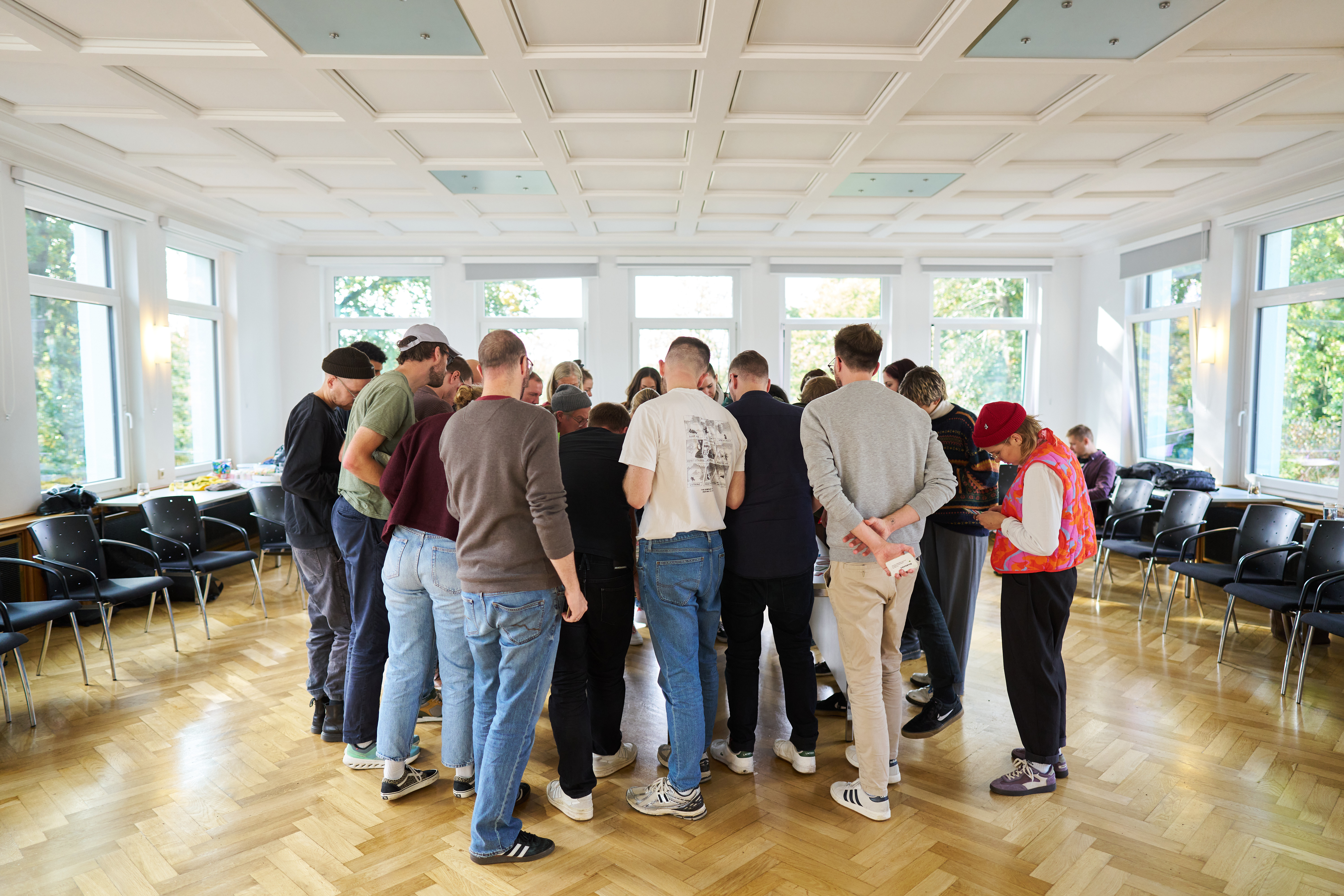 Eine Gruppe Menschen steht in einem Saal mit hellem Parkettboden. Sie bilden einen Kreis und schauen alle in die Mitte.