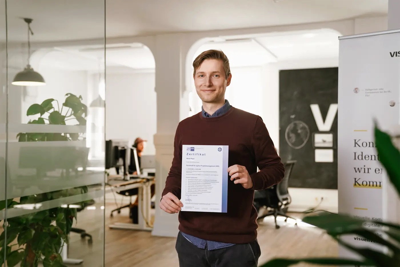 Lächelnd hält Rune ein Zertifikat der IHK vor sich in Händen. Er steht in einem Büroraum.