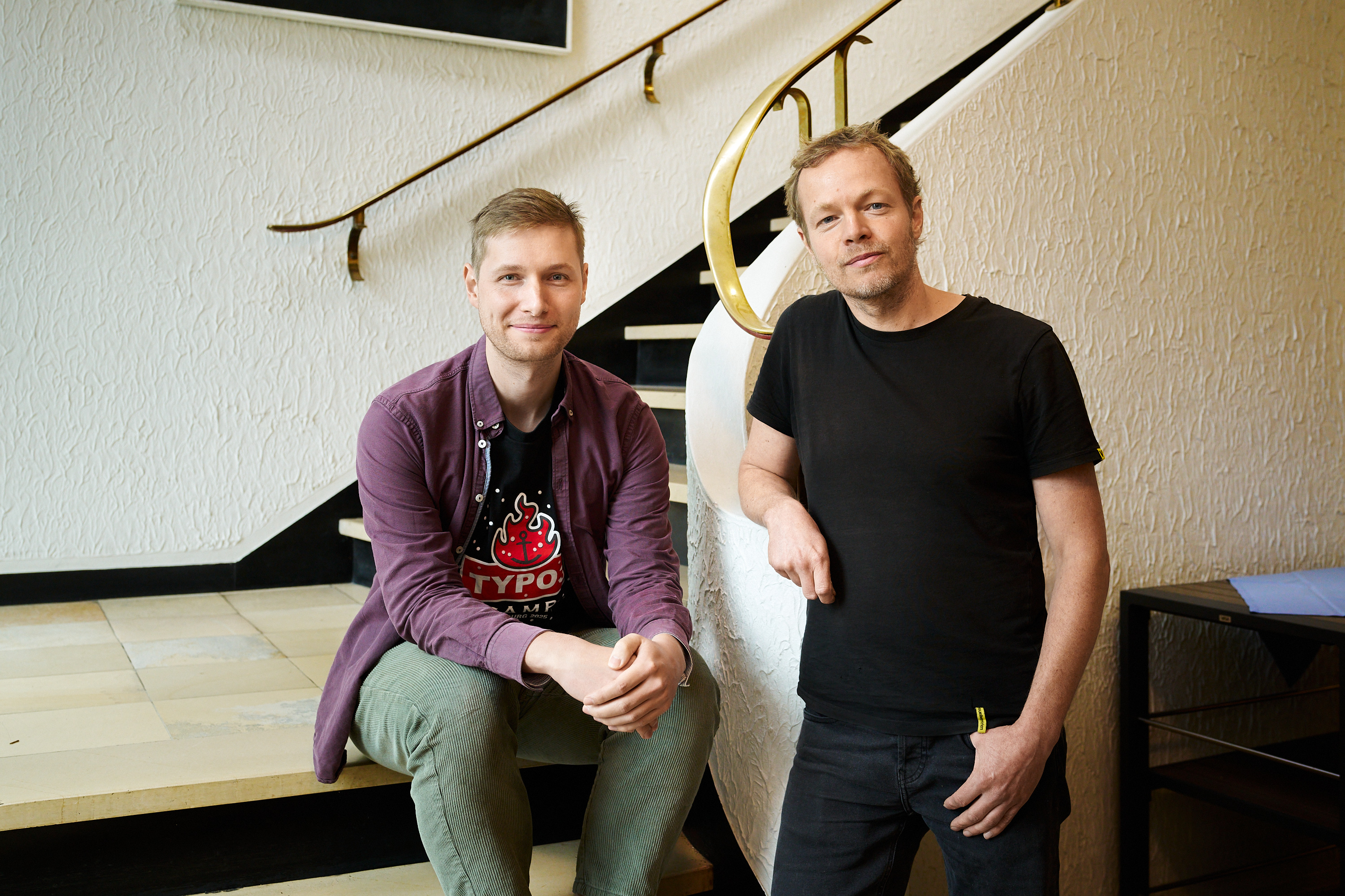 Rune und Timon an einem hellen Treppenaufgang. Rune sitzt auf der Treppe und trägt ein weinfarbenes, offenes Hemd und eine grüne Hose. Timon steht an das Geländer gelehnt. Er hat ein schwarzes T-Shirt und eine dunkelblaue Hose an.