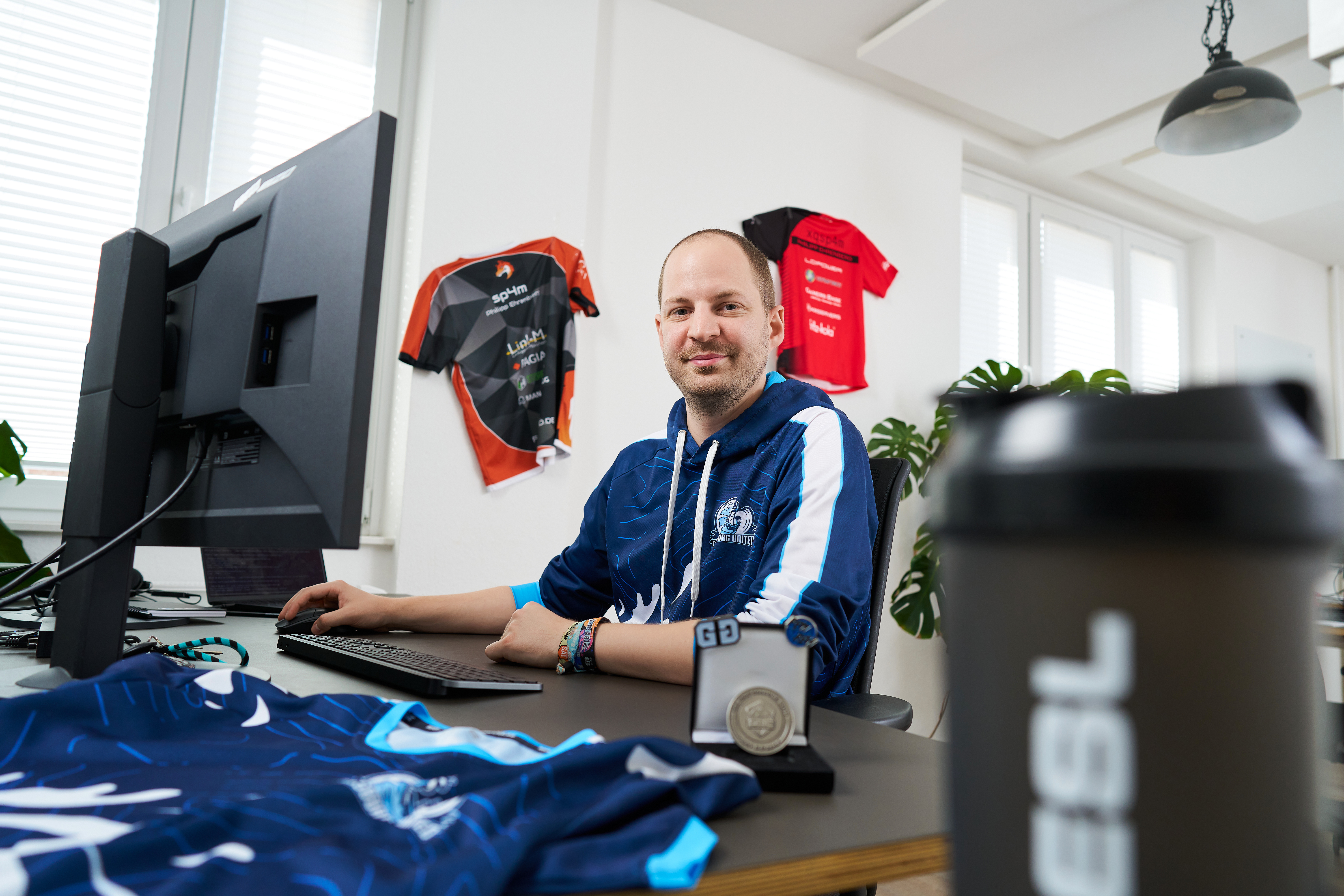 visuellverstehen-Mitarbeiter Philipp sitzt an seinem modernen, hellen Arbeitsplatz in der Agentur und lächelt direkt in die Kamera. Im Hintergrund hängen zwei E-Sport-Trikots an der Wand, ein weiteres liegt neben Philipps Monitor auf dem Schreibtisch. Im Vordergrund ist eine Sporttrinkflasche mit dem Aufdruck ESL zu sehen.