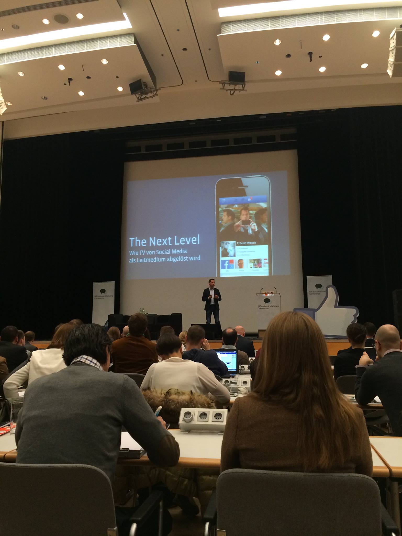 Viele Personen sitzen bei der Marketingkonferenz in einem Raum und sehen zu einer Person, die vorn etwas präsentiert.