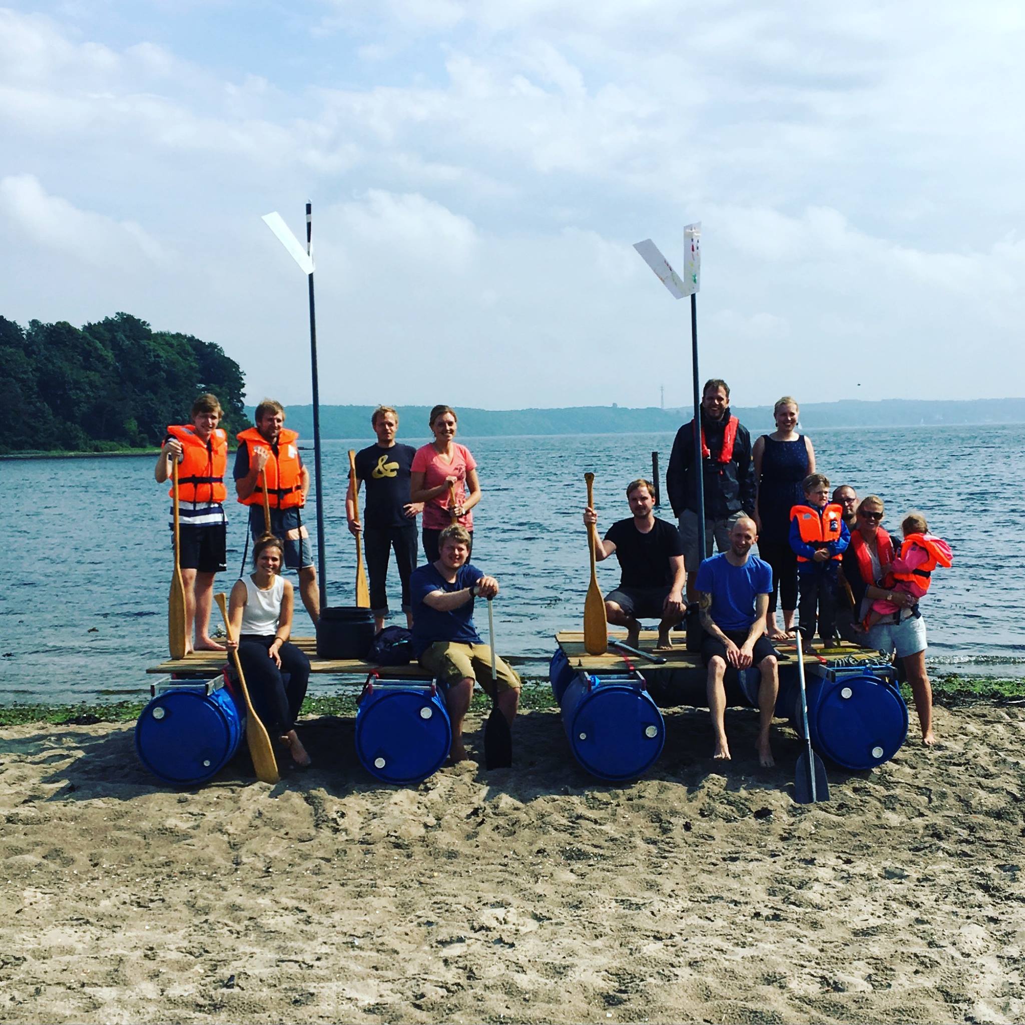 Mitarbeitende von visuellverstehen posieren am Strand auf und vor zwei selbstgebauten Flößen.