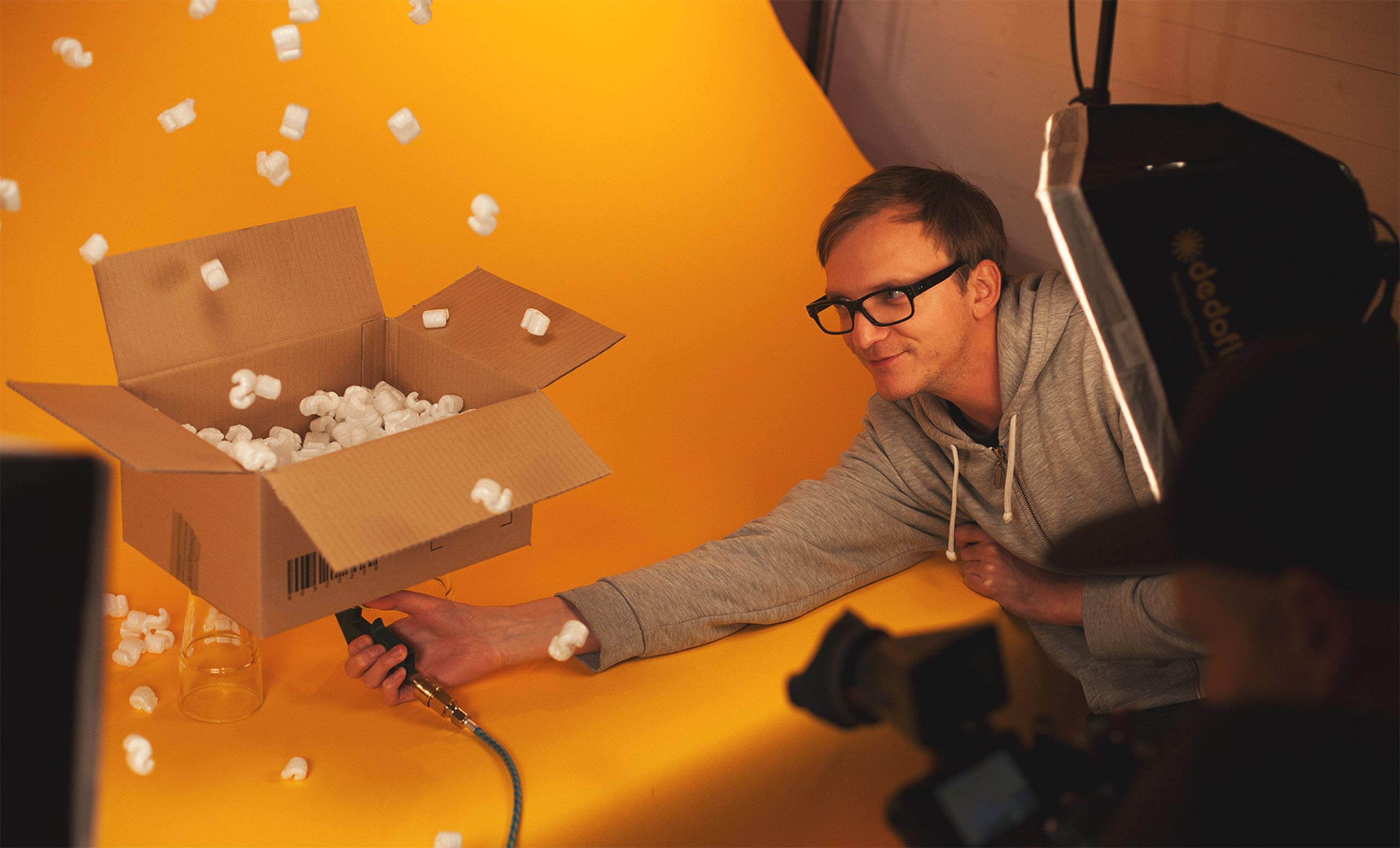 Foto vom Making-Of des vv-Films 2016: Ein offener Pappkarton voller Styropor-Bälle steht vor gelbem Hintergrund auf zwei Gläsern, ein Mitarbeiter reckt sich lächelnd nach dem Karton und hält ein Gerät an dessen Unterseite, sodass die Bälle in die Luft schweben.
